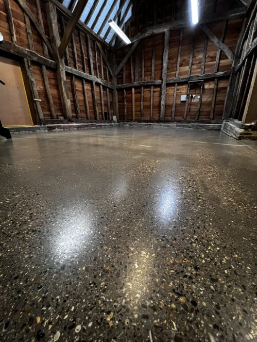 Polished concrete floors London shine inside a rustic wooden barn with exposed beams and wooden walls; overhead lights and daylight highlight the glossy, reflective surface.