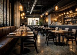 A modern, cosy restaurant with wooden tables, black chairs, warm hanging lights, and exposed brick walls. Tables are set with plates and glasses, and the bar area is visible in the background. No people are present.
