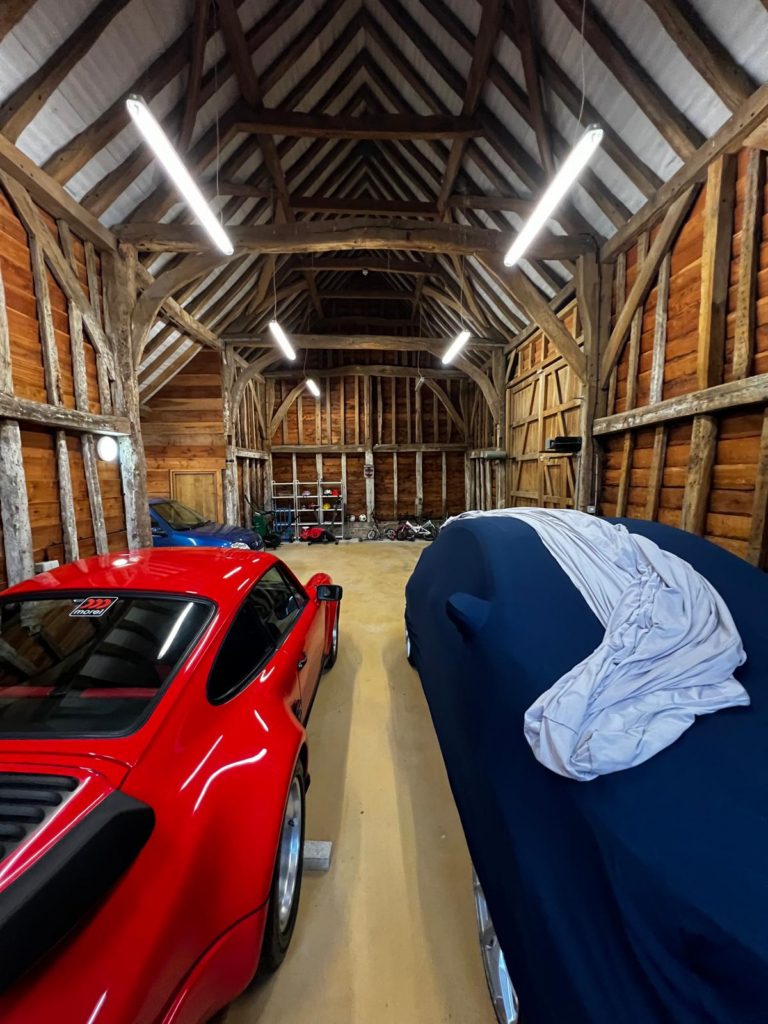 A spacious wooden barn with exposed beams, housing a red sports car and a covered vehicle. Shelving with various items is visible at the back, and overhead fluorescent lights illuminate the space.