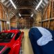 A spacious wooden barn with exposed beams, housing a red sports car and a covered vehicle. Shelving with various items is visible at the back, and overhead fluorescent lights illuminate the space.