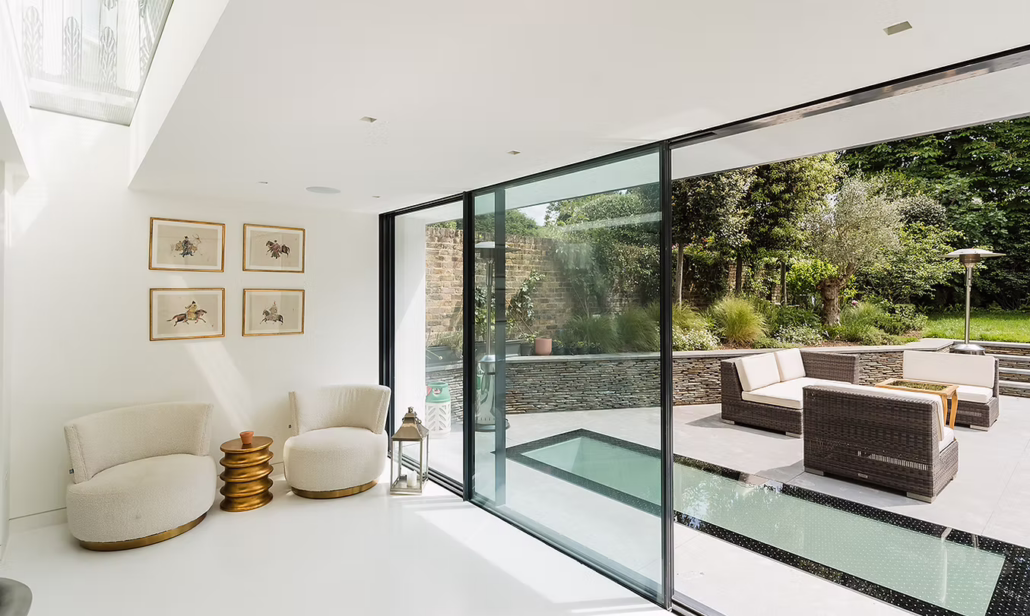 Modern living room with two cream armchairs, gold nesting tables, and framed artwork on white walls. Large glass sliding doors open to a patio with outdoor seating and a landscaped garden.