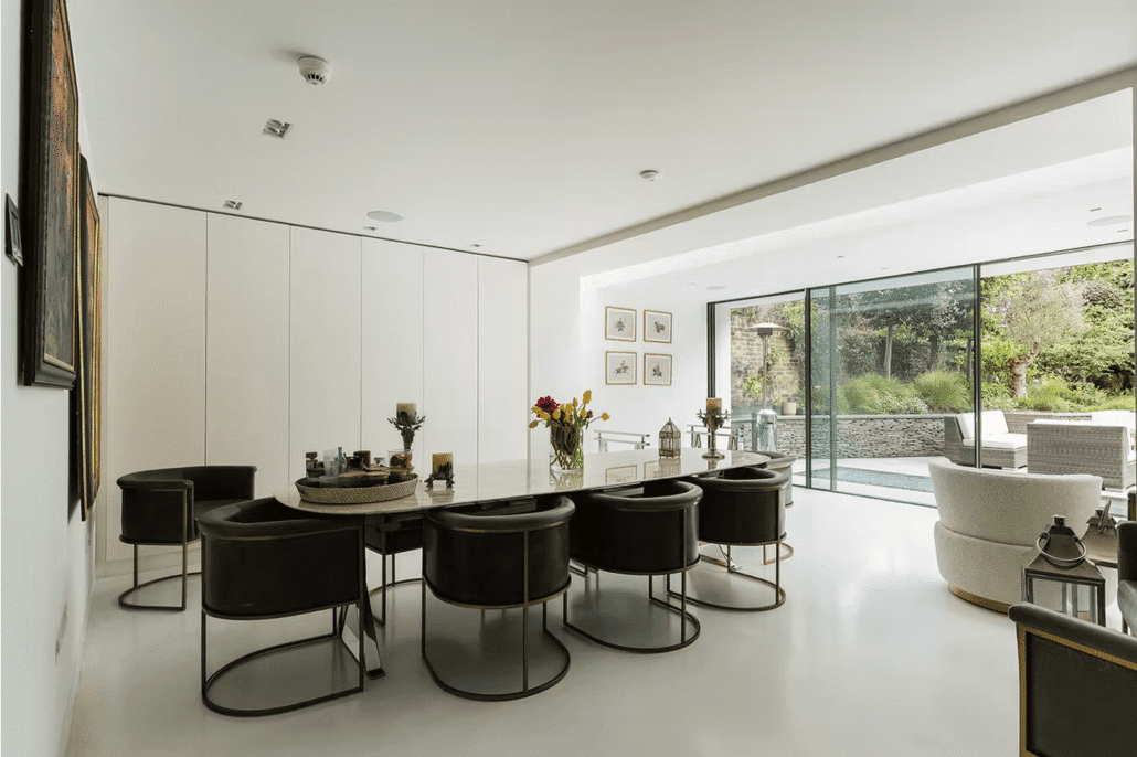Modern dining room with a round table surrounded by black chairs, white walls, large windows overlooking a garden, and contemporary decor with flowers and framed art. Ample natural light fills the space.