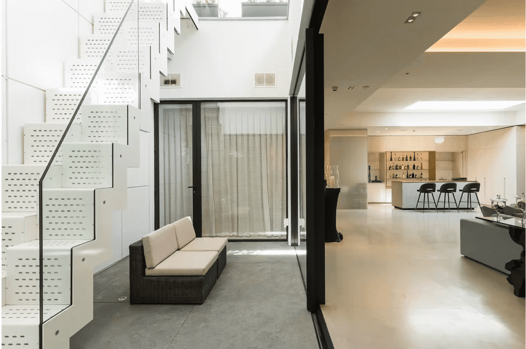 A modern, minimalist interior with white perforated metal stairs, glass balustrade, a wicker loveseat, concrete floors, and an open living area featuring a kitchen bar with black stools and neutral furnishings.