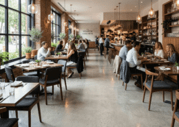 A modern, stylish restaurant with large windows, wooden tables, potted plants, and people dining in pairs and groups. The open kitchen is visible in the background, and natural light fills the space.