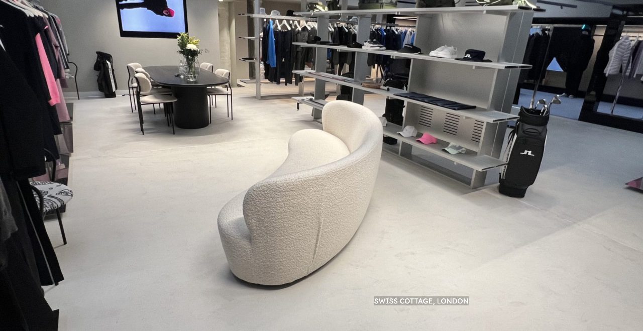 A modern clothes shop with a curved white microcement sofa in the centre, shelves of folded clothes, rails of hanging garments, a golf bag, and a round table with flowers in a vase. A label reads SWISS COTTAGE, LONDON.