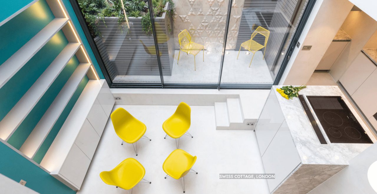 Modern, minimalist kitchen and dining area with white microcement floors, marble worktops, four yellow chairs, and floor-to-ceiling windows opening to a small patio with two more yellow chairs. Shelves and teal feature wall visible.