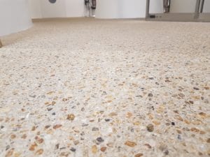 Close-up view of a polished concrete floor with exposed aggregate stones from the LPC Polished Concrete Portfolio; blurred background shows part of a modern indoor space with white walls and some furniture.