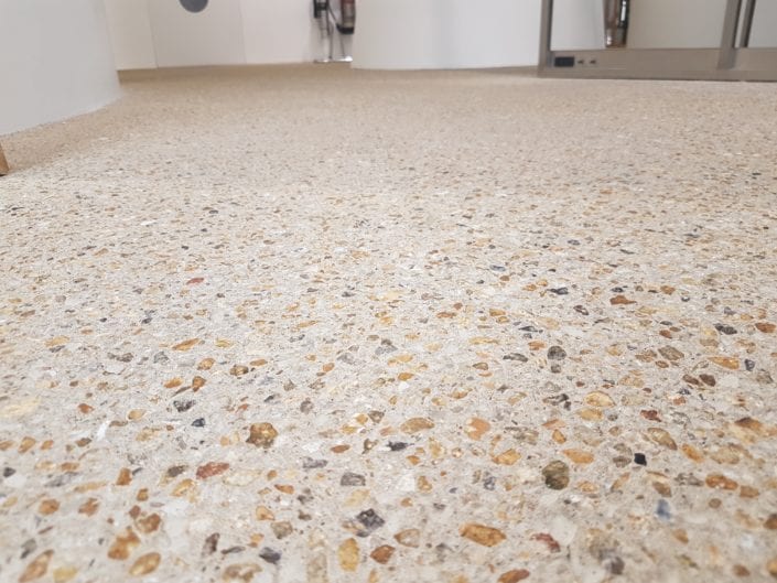 Close-up view of a polished concrete floor with exposed aggregate stones from the LPC Polished Concrete Portfolio; blurred background shows part of a modern indoor space with white walls and some furniture.