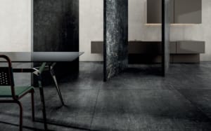 A modern minimalist kitchen with dark textured flooring and wall panels, floating cupboards, a glass-topped dining table, and a chair with a green seat. The sleek, industrial design showcases London Polished Concrete’s products and services for a refined finish.