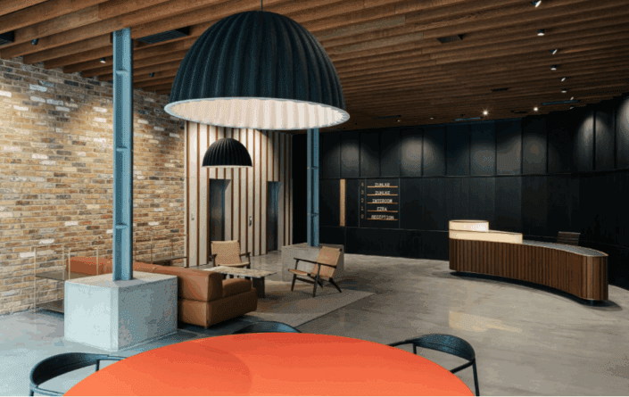 Modern office lobby with a curved wooden reception desk, brown leather seating area, exposed brick wall, and polished concrete floors. Large black pendant lights and a timber slat ceiling add style, while a round orange table stands in the foreground. London-inspired design.