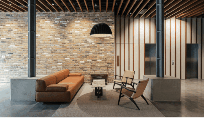 Modern lobby with a brown leather sofa, two woven chairs, a small coffee table, a large black pendant light, brick feature wall, polished concrete floors, and two lifts set in a minimalist, industrial-style London interior.