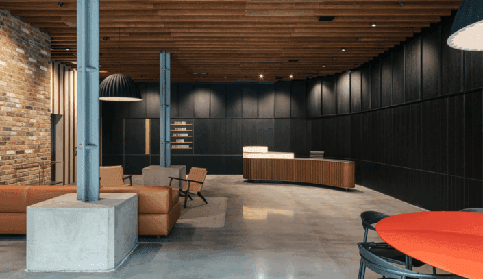 Modern office lobby with a sleek wooden reception desk, black panel walls, exposed brickwork, brown and black furniture, a red table, polished concrete floors London, and a wooden slat ceiling with pendant lights.