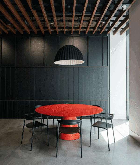 A modern dining area with a round red table, four black chairs, a large black pendant lamp, exposed wooden ceiling beams, and dark vertical wall panels sits atop polished concrete floors in London, flooded with natural light from a window.