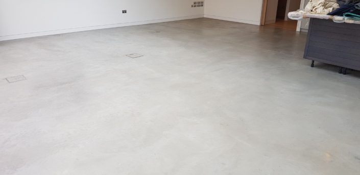 A large room with smooth, light grey polished concrete flooring—reflecting the latest Polished Concrete Cost in London (2026) trends—white walls, and minimal furniture; several floor access covers are visible.