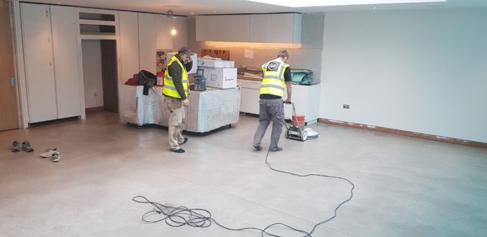 Two workers in yellow safety vests use a floor polishing machine on concrete floors. Nearby boxes and equipment hint at ongoing work. Curious about Polished Concrete Cost in London (2026)? Check our Pricing Guide & Advice for details.