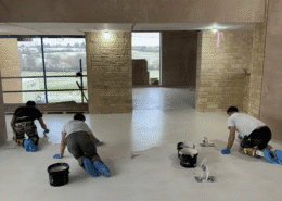 Three people wearing gloves and shoe covers are kneeling on a floor, applying a white coating in a large, unfinished room with exposed brick walls and a big window—showcasing the process involved in polished concrete vs other flooring finishes. Paint buckets and rollers are nearby.