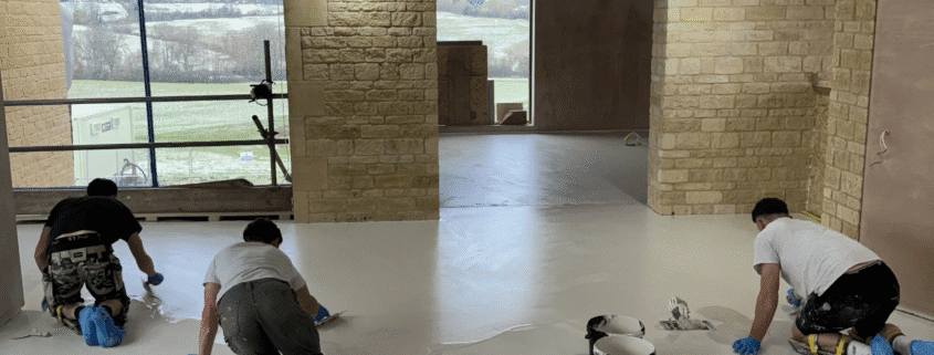 Three people wearing gloves and shoe covers are kneeling on a floor, applying a white coating in a large, unfinished room with exposed brick walls and a big window—showcasing the process involved in polished concrete vs other flooring finishes. Paint buckets and rollers are nearby.