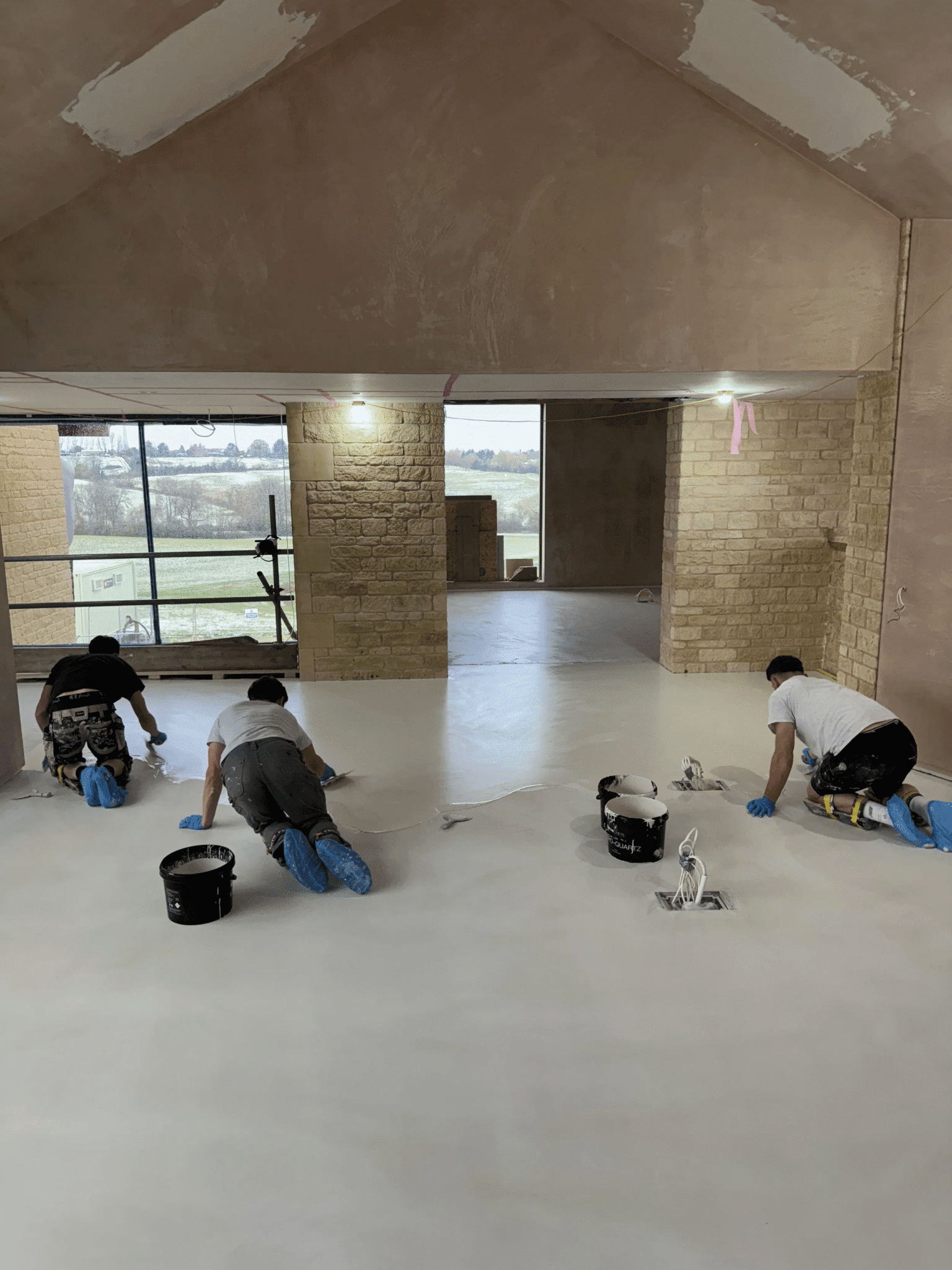 Three people wearing gloves and shoe covers are kneeling on a floor, applying a white coating in a large, unfinished room with exposed brick walls and a big window—showcasing the process involved in polished concrete vs other flooring finishes. Paint buckets and rollers are nearby.
