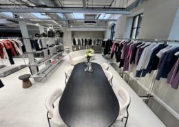 A modern showroom in London with polished concrete floors, featuring a long black table and six chairs, a white sofa, and rails displaying neatly arranged clothing along the bright, minimalist space with large windows.