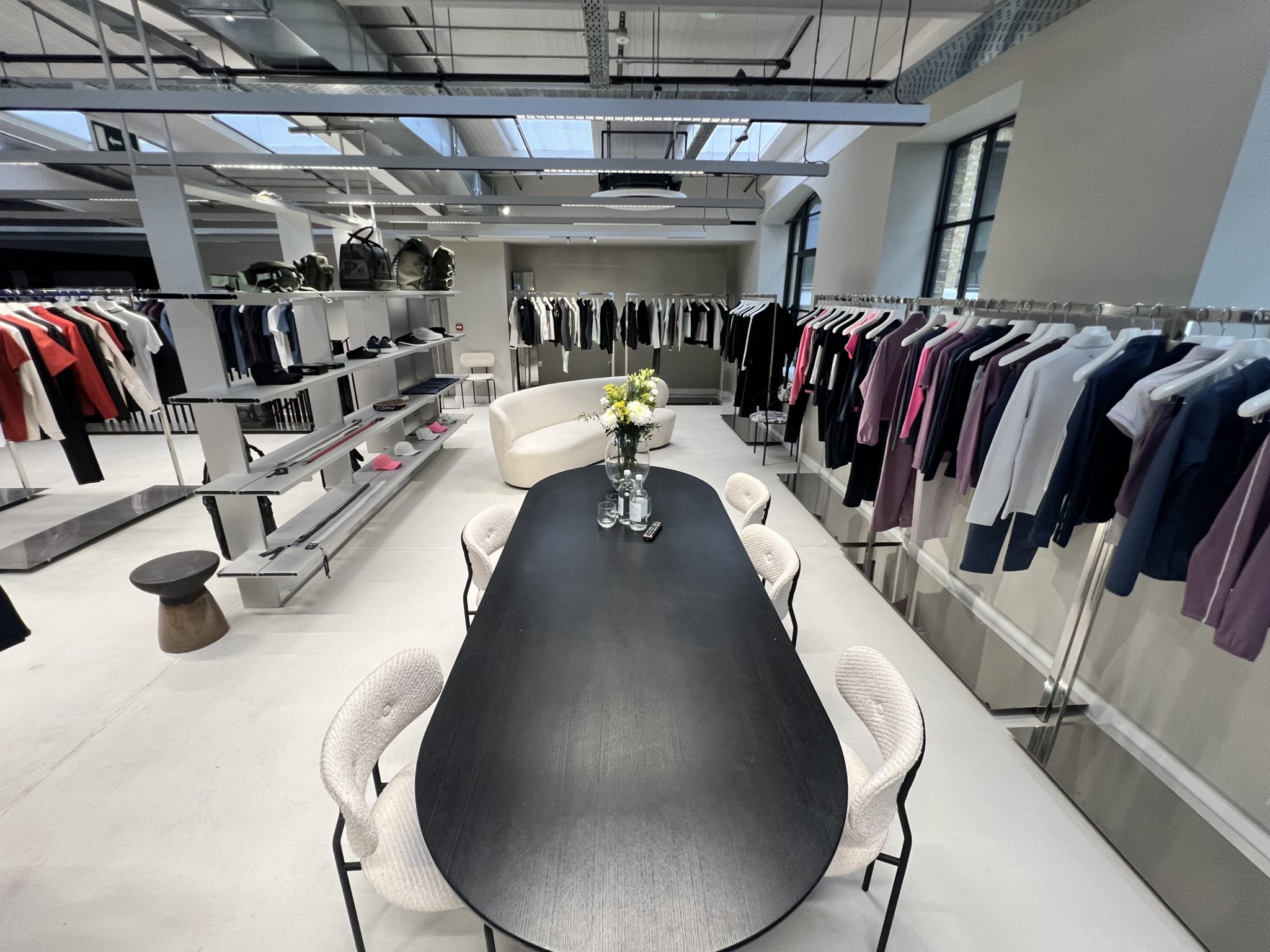 A modern showroom in London with polished concrete floors, featuring a long black table and six chairs, a white sofa, and rails displaying neatly arranged clothing along the bright, minimalist space with large windows.