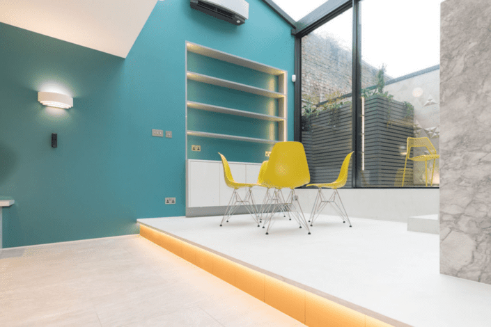 A modern dining area with four yellow chairs around a small white table, teal feature wall with built-in shelving, and a raised floor with under-lighting, featuring London Polished Concrete’s products and services for a sleek urban feel.