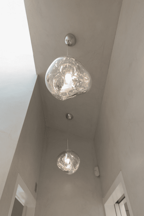 Two modern pendant lights with clear, organic-shaped glass shades hang from a high, light grey ceiling in a minimalistic hallway featuring London Polished Concrete’s products and services. Natural light enters from a skylight above.