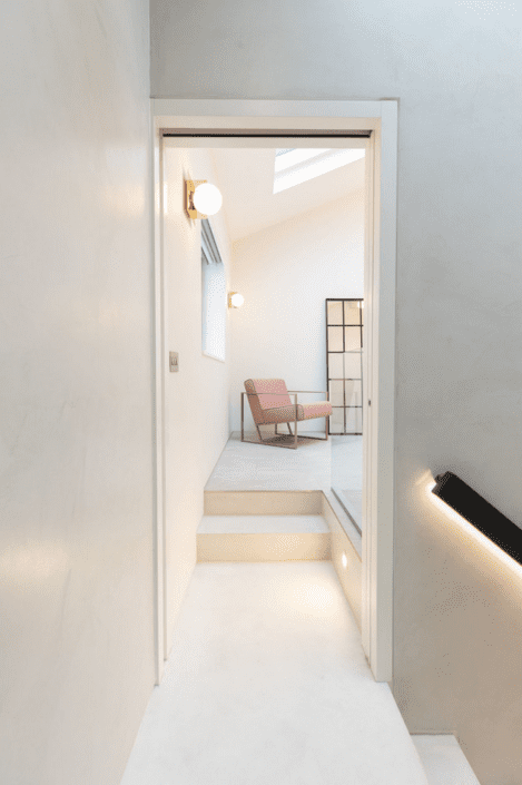 A minimalist, bright hallway leads to a sunlit room with steps. Featuring London Polished Concrete’s Products and Services, a pink armchair sits near a black-framed folding screen, with round wall lights warming the modern, white interior.