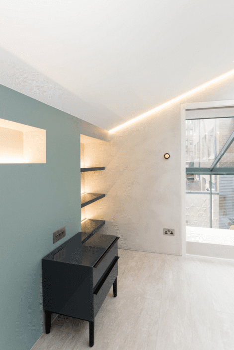 Modern minimalist room with teal feature wall, built-in illuminated shelves, black glossy chest of drawers, and large window, featuring light-coloured flooring by London Polished Concrete’s Products and Services under a sloped ceiling with recessed lighting.