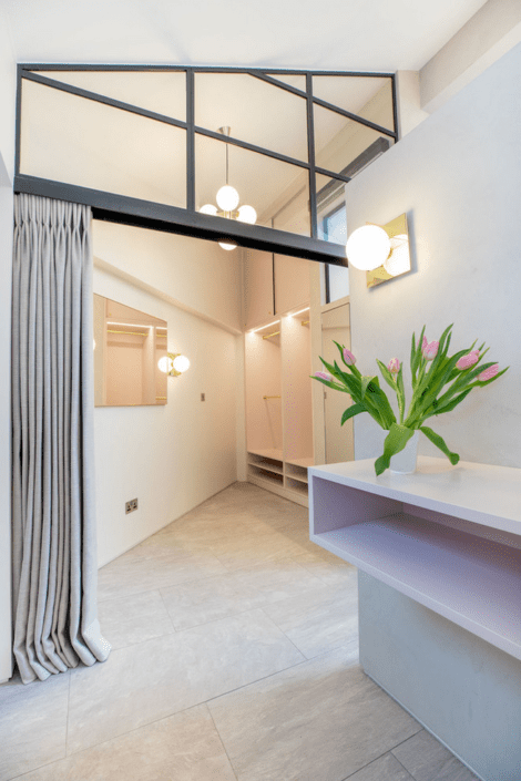 A modern, minimalist hallway with light-coloured walls and floor tiles crafted using London Polished Concrete's Products and Services, a hanging pendant light, a grey curtain, a pink floating shelf holding tulips, and wall mirrors reflecting the lighting.