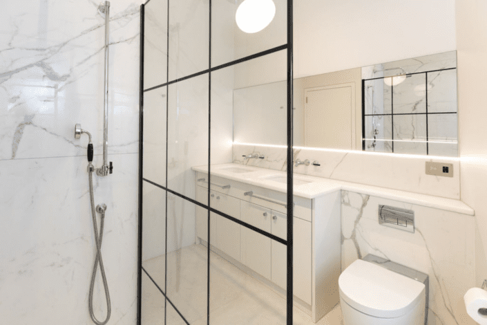 A modern bathroom with white marble walls, a glass shower screen with black grid lines, double-sink vanity, wall mirror, built-in lighting, and a toilet. London Polished Concrete’s products and services enhance the clean, minimalist space.