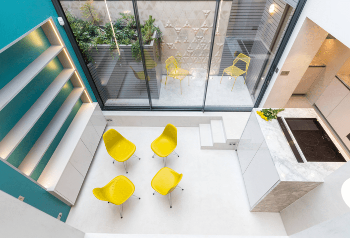 A modern, minimal kitchen and sitting area features London Polished Concrete’s products and services, with four bright yellow chairs around a white floor, glass doors to a patio with two yellow chairs, and built-in shelving on a turquoise wall.