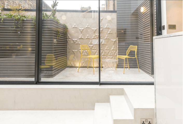 A small modern outdoor patio features two yellow chairs and a clear round table, with geometric-patterned walls crafted using London Polished Concrete’s Products and Services, potted plants, and a glass enclosure providing a view from inside.