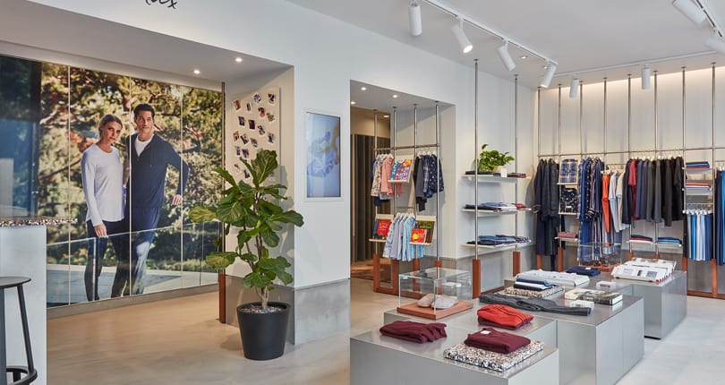 Knightsbridge MC Modern clothing shop interior with neatly folded clothes on metal tables, racks of hanging garments, a large plant, and a wall mural of two people outdoors. The bright space features minimalistic decor and stylish microcement floors London.