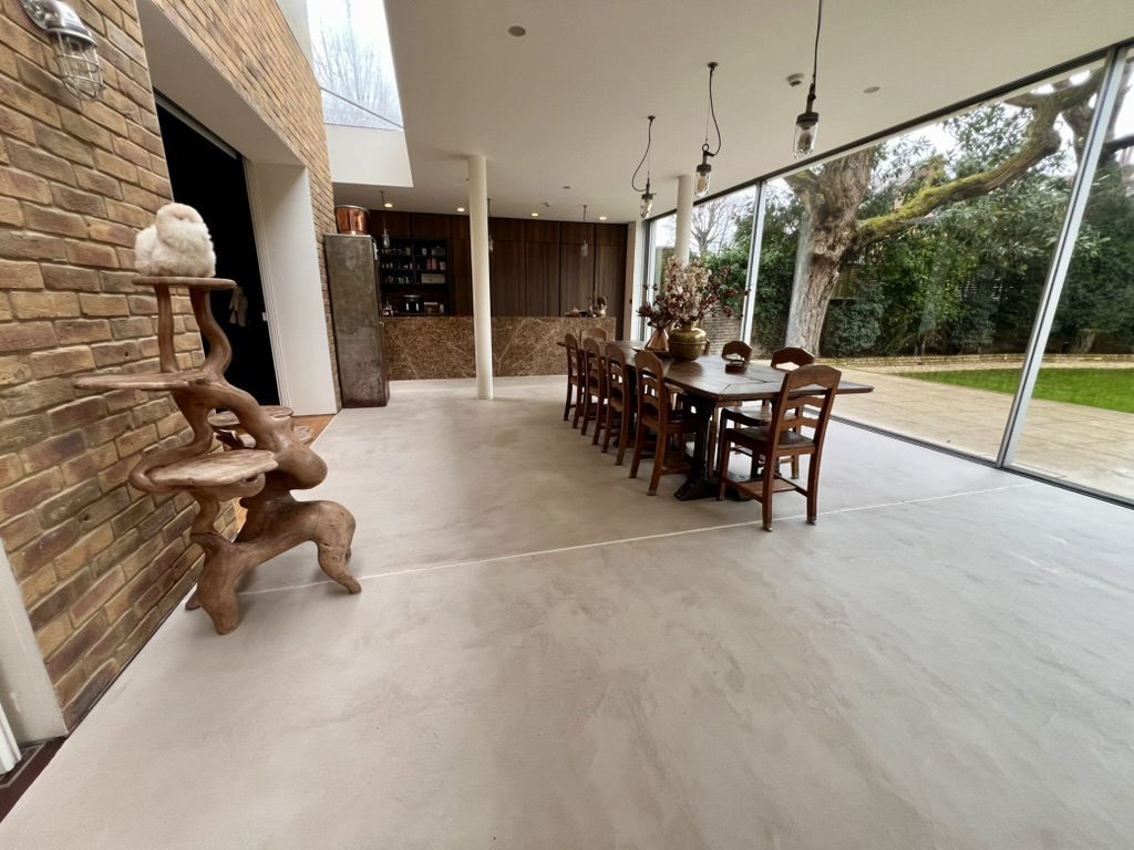 A modern dining area with large glass walls, a long wooden table with chairs, and a unique wooden cat tree with a cat sitting on top; outside, there is greenery and a stone patio.