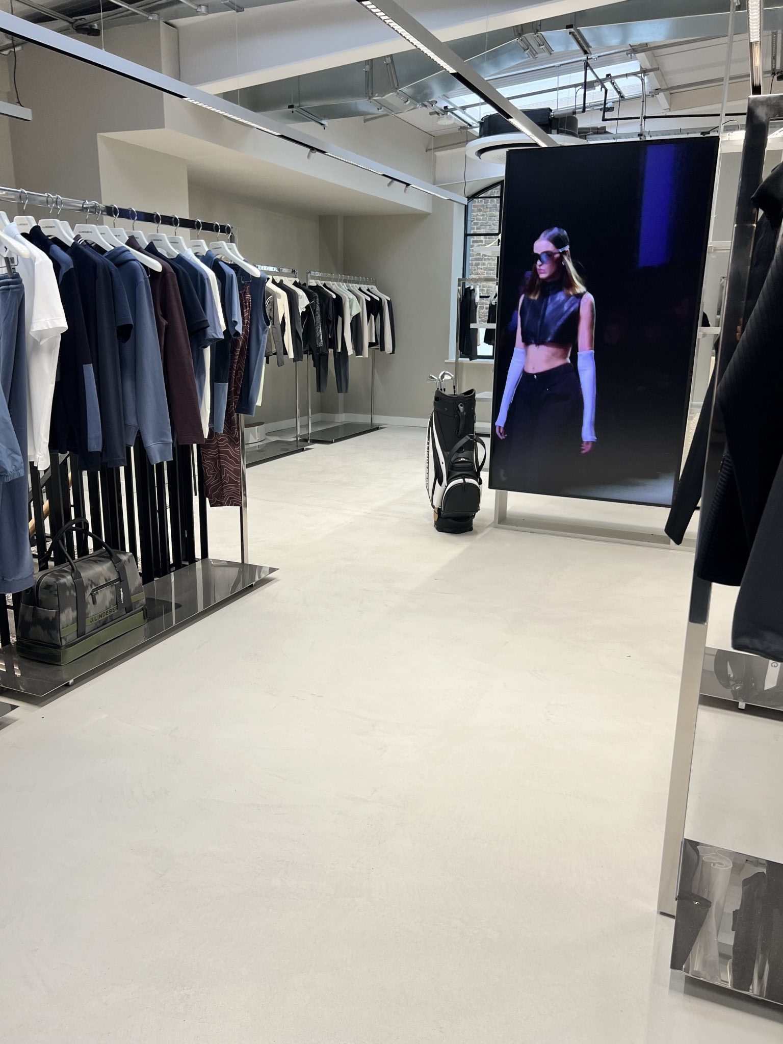 A modern clothes shop interior with rails of neatly arranged clothes, a golf bag, and a large digital screen displaying a model in a black crop top and sunglasses walking on a catwalk.