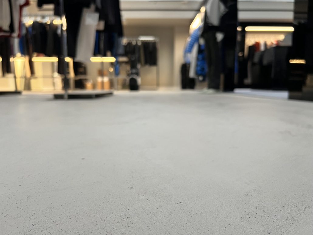 A low-angle view of a clothes shop interior with rails of clothes and bright lighting, focusing on the smooth, light-coloured floor in the foreground.