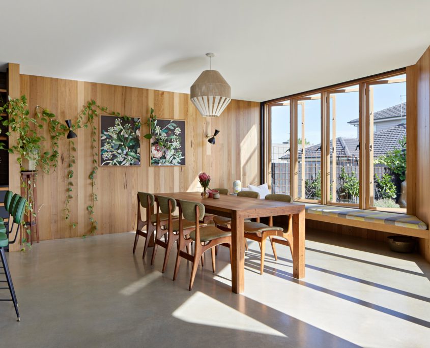 Bright dining area with wooden walls and large windows, featuring a wooden table with six green chairs, a built-in window seat, hanging plants, and framed artwork—showcasing polished concrete versus other flooring finishes as sunlight streams in.