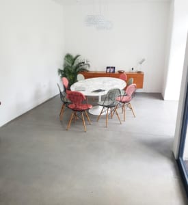A modern dining room with a round marble table, six colourful chairs, a plant in the corner, and a wooden sideboard with books and a lamp—all set against white walls and a concrete floor finished using London Polished Concrete's products and services.