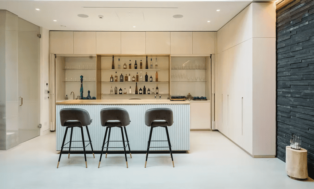 A modern home bar with a light wood and white colour scheme, three bar stools at a central island, glass shelves with bottles and glasses, and sleek minimalist cabinetry.