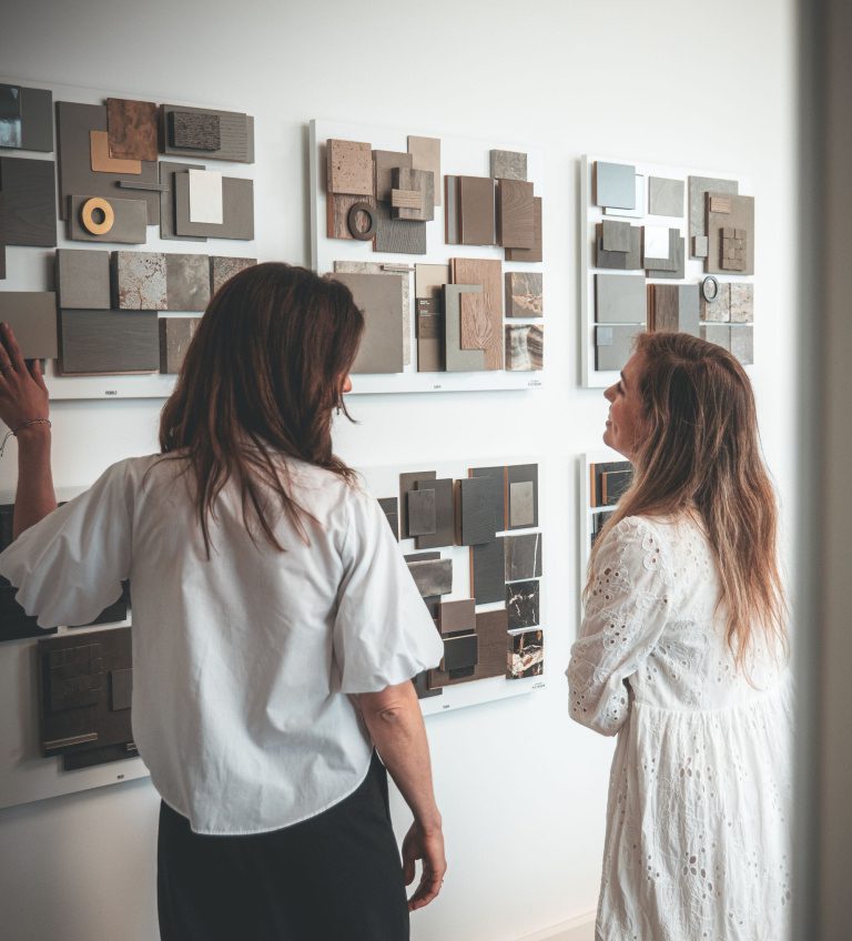 Two women stand in front of a wall displaying several abstract art pieces—layered, rectangular works in neutral tones—whilst discussing or observing the artworks closely, much like comparing polished concrete versus other flooring finishes.