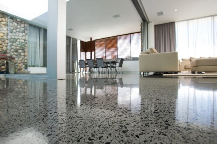 A close-up view of a polished microcement floor in a modern, open-plan living space with large windows, a dining table and chairs, a beige sofa, and stone wall accents—showcasing the elegance of microcement London style.