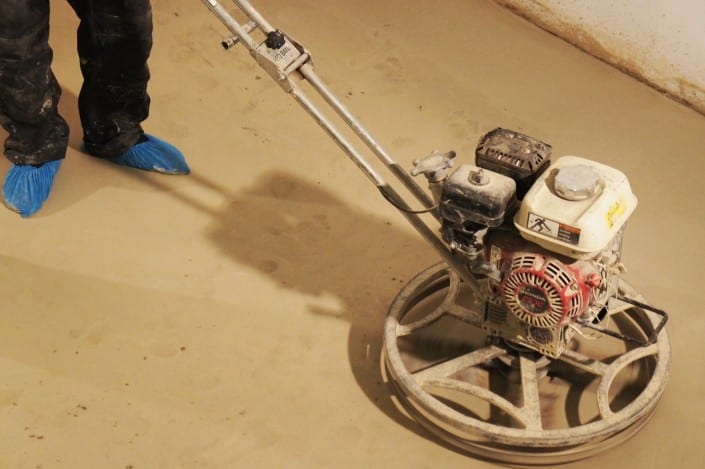 SONY DSC A person wearing blue shoe covers operates a power trowel machine to smooth a wet concrete floor, preparing the surface for stunning microcement London finishes in an indoor setting.