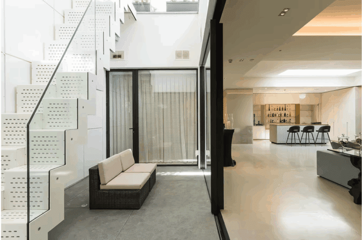 Modern interior with white perforated metal staircase, glass balustrade, beige cushioned bench, and an open-plan living area featuring a kitchen with a breakfast bar and stools, neutral tones, polished concrete floors London, and minimalist decor.