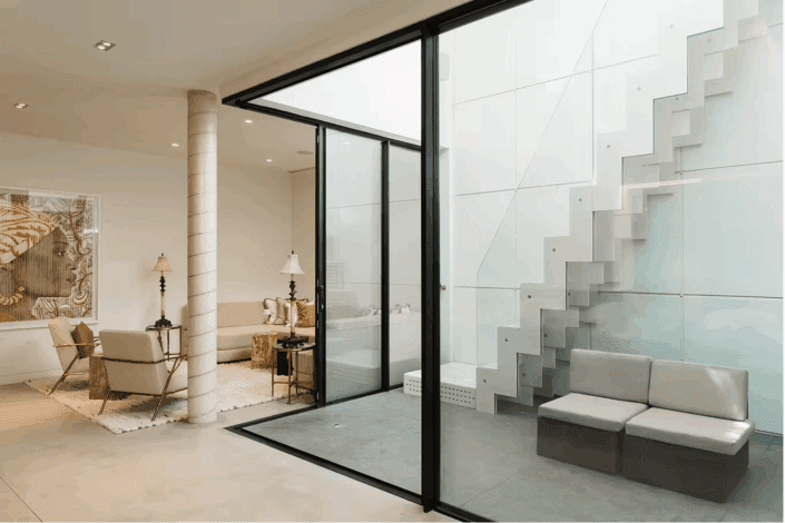 Modern indoor space with polished concrete floors London, where a glass wall separates a lounge area from a minimalist patio. The patio features grey seating and a staircase with glass panel railings, while the lounge offers beige armchairs and a decorative rug.