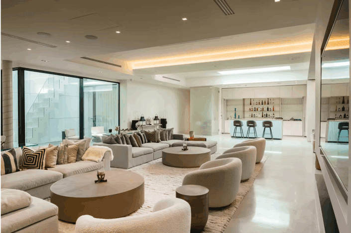 A spacious, modern living room with large grey sofas, round coffee tables, curved beige chairs, and a plush rug sits atop polished concrete floors. A sleek bar area with stools stands in the background, while floor-to-ceiling windows fill the space with natural light.