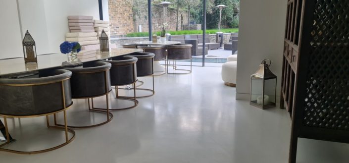A modern, minimalist room with round black chairs around a white dining table, polished concrete floors in London style, large glass doors opening to a patio with stacked towels and decorative lanterns on the floor and table. Gardens are visible outside.