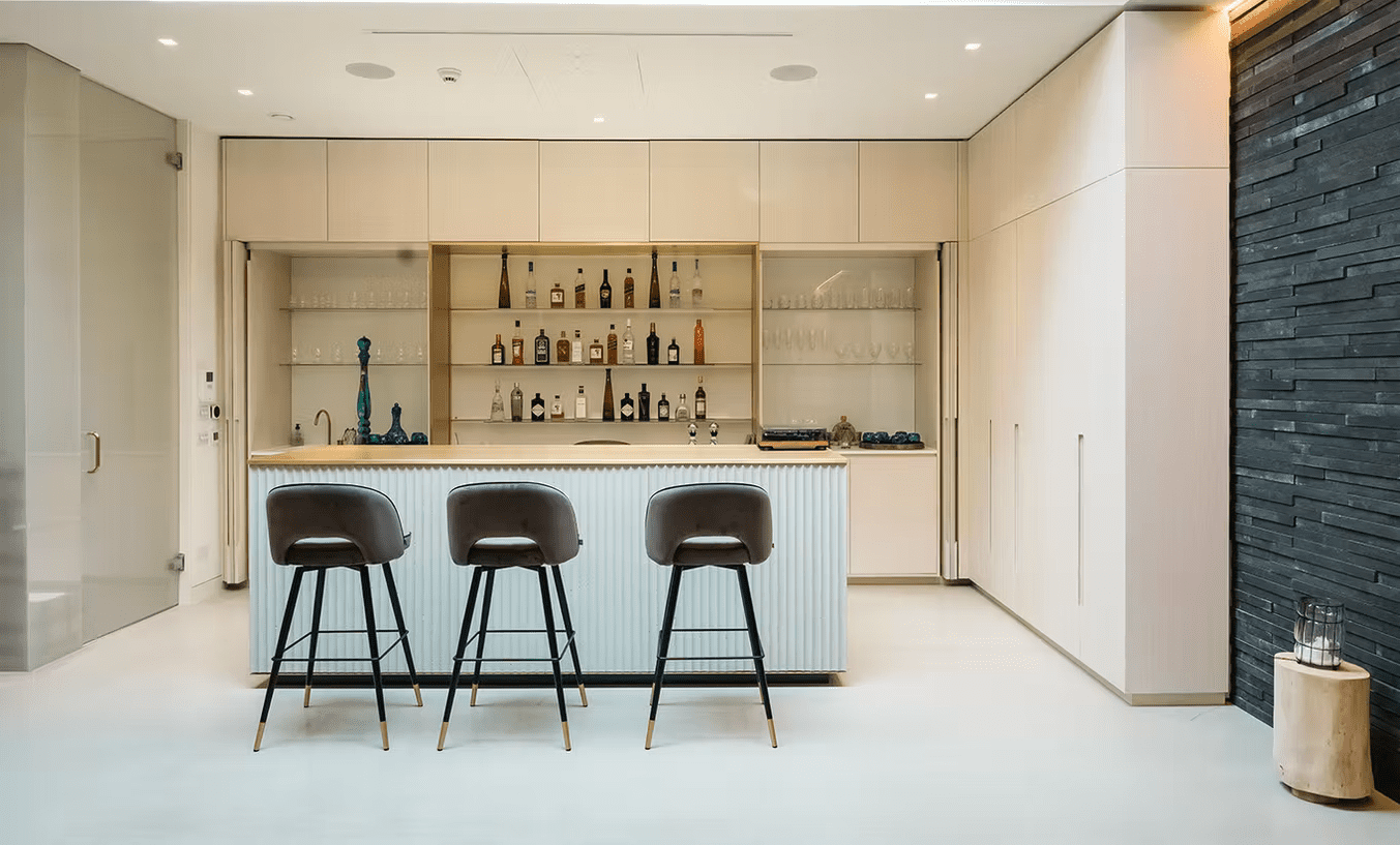 Modern home bar with a sleek island, three bar stools, built-in shelves stocked with bottles and glasses, light wood cabinetry, and a dark stone feature wall on the right. Microcement London flooring enhances the space’s clean and minimalist vibe.