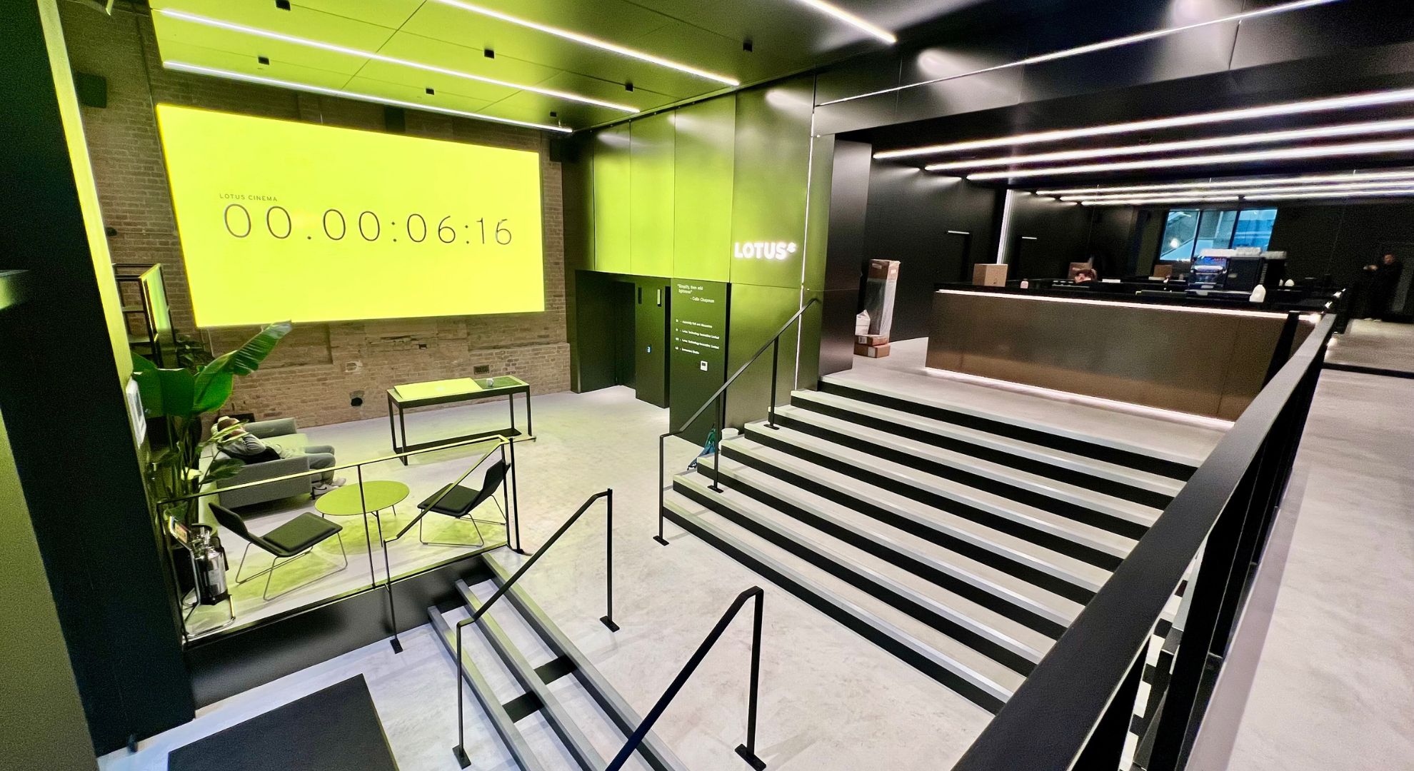 A modern lobby with black and neon yellow-green accents, featuring microcement London finishes, a reception desk, staircase, lounge seating, and a large screen displaying a countdown timer and the word LOTUS on the wall.